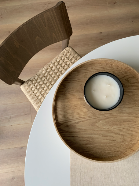 Mesa blanca con bandeja de madera y vela en un recipiente negro, junto a una silla.